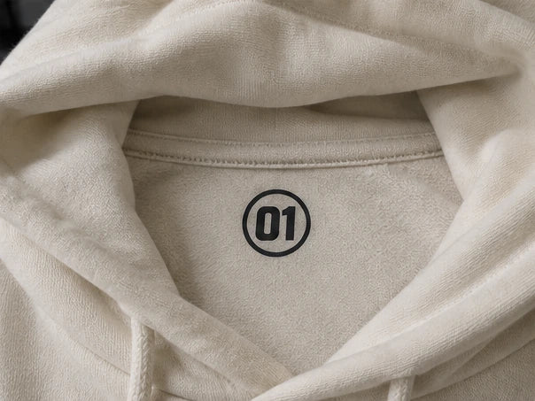 Ivory hoodie with ONE front artwork, PIT CREW sleeve lettering, and an 01 neck label. — Inside-neck application